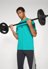 Turkise træningst-shirt med sort tre-stribedesign på ærmerne, båret af en person, der løfter en vægtstang, parret med sorte bukser.