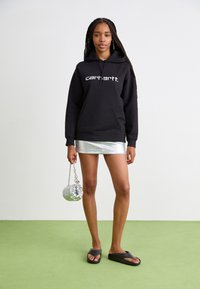 Felpa con cappuccio nera con logo "carhartt." bianco, abbinata a una mini gonna argento e sandali grigi. Tiene in mano una borsa rotonda metallica.