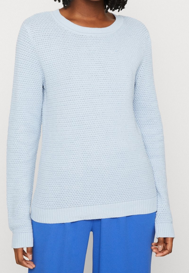 Femme portant un pull tricoté texturé bleu clair avec un col rond et des manches longues, associé à un pantalon bleu vif.
