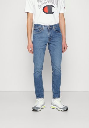 Personne portant un jean slim bleu, un t-shirt blanc Champion et des baskets blanches avec des accents jaune néon, debout devant un fond blanc uni.