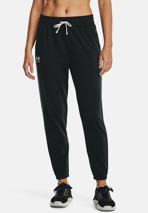Femme portant un pantalon de jogging Under Armour noir avec un cordon blanc et des chaussures de sport noires, debout devant un fond uni.