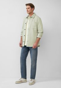 Lichtgroene button-up jas, lichtgele shirt, blauwe jeans en crèmekleurige sneakers. De jas heeft een zak en opgerolde mouwen, casual pasvorm.