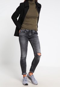 Abrigo negro, jersey de cuello alto oliva, jeans pitillo gris desgarrados y zapatillas gris claro. Nota: desgarro visible en la rodilla y puños doblados.