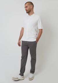 Korte mouwen witte gebreide polo met een gestructureerd diamantpatroon, gecombineerd met grijze broek en witte sneakers, gedragen door een staande mannelijke model.