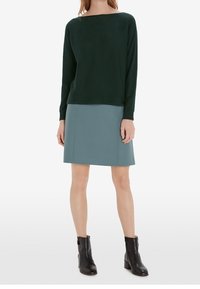 Donna che indossa un top a maniche lunghe verde scuro, una gonna blu al ginocchio e stivali neri alla caviglia, in piedi davanti a uno sfondo bianco.