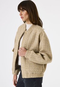 Femme portant une veste texturée beige sur un haut blanc et un jean sombre, se tenant de côté contre un fond blanc uni.