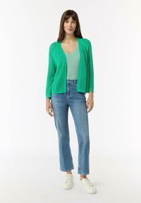 Grüne, gestrickte Strickjacke über einem gestreiften Tanktop, kombiniert mit hellblauen, geraden Jeans und weißen Sneakers. Einfaches Design, lässiges Erscheinungsbild.