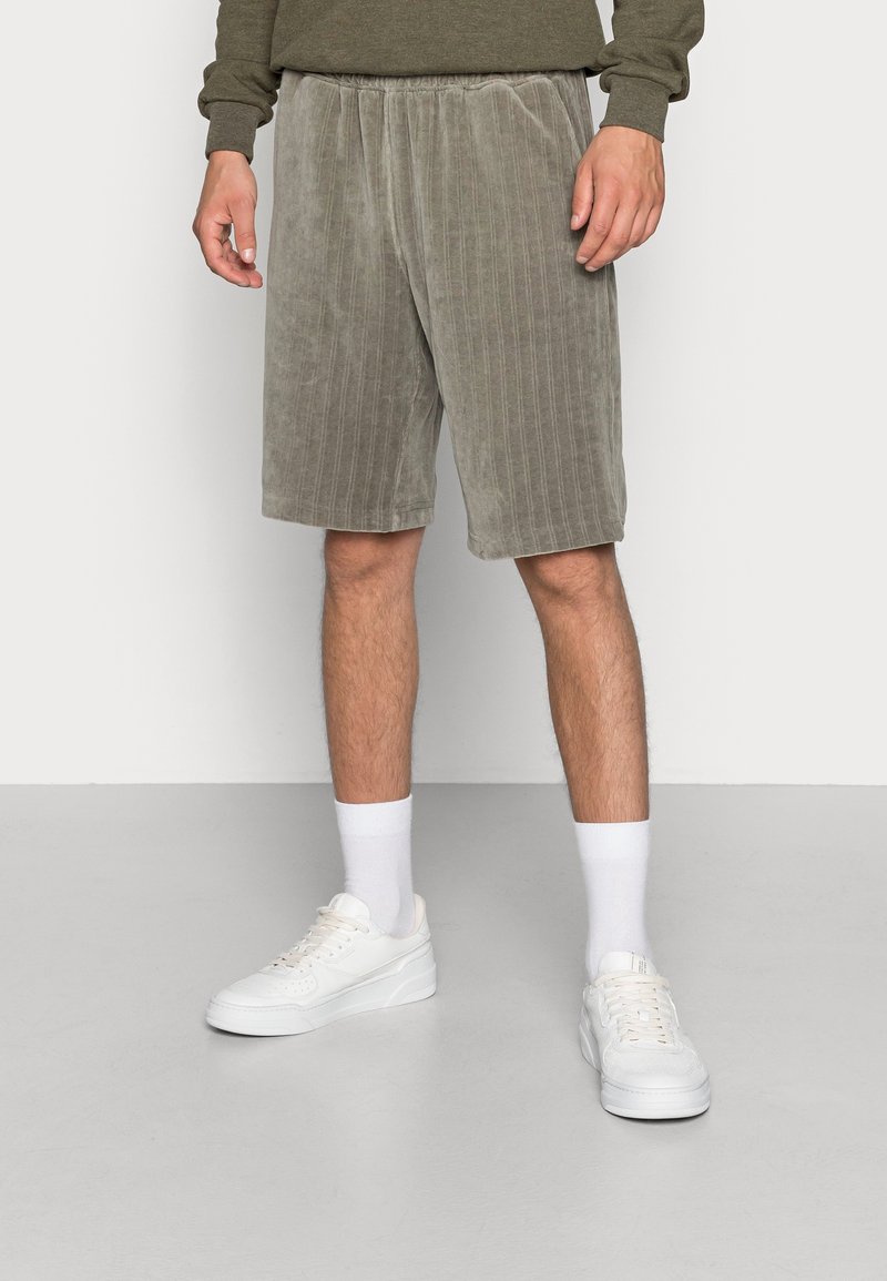 Homme portant un short en velours couleur taupe à rayures verticales, des chaussettes blanches, des baskets blanches, et un haut à manches longues vert olive.