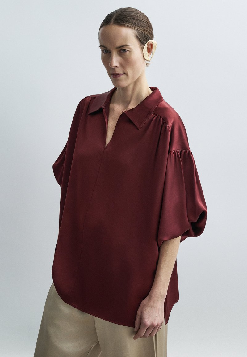 Kvinde iført en løs vinrød bluse med puffede korte ærmer og beige bukser, stående mod en ensartet lysegrå baggrund.