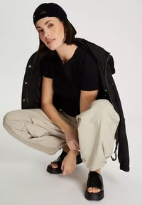 Czarny T-shirt, beżowe luźne spodnie, czarne sandały na płaskiej podeszwie. Stylizowana kurtka zarzucona na ramię, krótkie włosy, w ciemnej czapce.