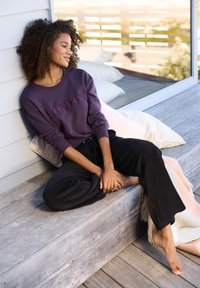 Lila Sweatshirt mit roter Stickerei "LONDON", kombiniert mit schwarzen gerippten Hosen. Modell sitzt barfuß auf einer Holzterrasse, mit weichen Kissen in der Nähe.