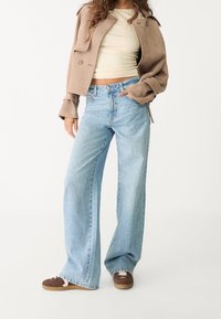 Jeans de pierna ancha en azul claro con cintura alta, que presentan un sutil patrón moteado y dobladillo deshilachado, combinados con un top corto beige y zapatos marrones.
