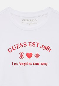 Λευκό μπλουζάκι με κόκκινο κείμενο που γράφει «GUESS EST. 1981», κόκκινη καρδιά και σύμβολα, και «Los Angeles 1201-1203» κάτω από το γιακά.