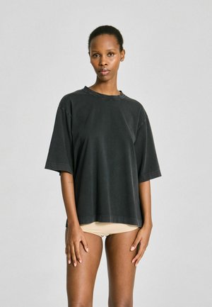 Femme debout, portant un t-shirt noir oversize et des sous-vêtements beiges, de face devant un fond uni gris clair.