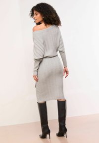 Graues Strickkleid mit schulterfreiem Design, gerippter Textur und taillierter Silhouette. Knielange schwarze Stiefel mit Absatz ergänzen das Outfit.