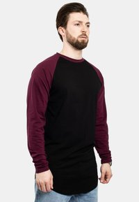 Duga majica s dugim rukavima koja ima crno tijelo i maroon raglan rukave, izrađena od pamučne tkanine s okruglim izrezom i zakrivljenim rubom.