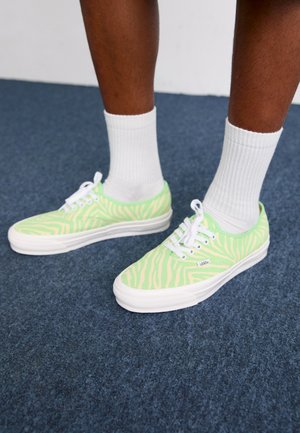 Zöld zebra mintás vászon tornacipők fehér gumitalppal és fűzőkkel, fehér bokazoknival viselve kék szőnyegfelületen.