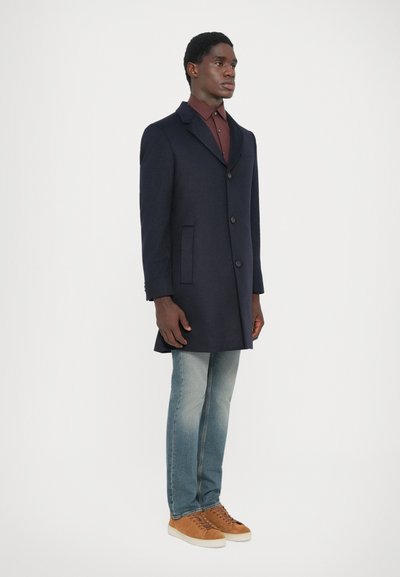 Marineblauer Wollmantel mit taillierter Form, mit Reverskragen und drei Knöpfen. Helle Jeans und beige Sneakers vervollständigen das Outfit.