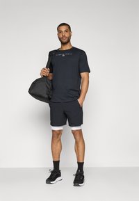 Schwarzes sportliches T-Shirt mit Logo, kombiniert mit dunklen Shorts mit weißer Borte. Das Modell hält eine schwarze Sporttasche und trägt schwarze Sneakers und Socken.