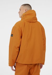 Narandžasta vodootporna jakna s kapuljačom, s opuštenim krojem i logotipom na rukavu. Glatka tekstura, minimalistički dizajn.