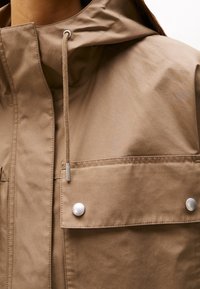 Lichtbruin jack met capuchon, voorzien van een koordsluiting, zilverkleurige drukknoopjes en een borstzak voor veilige opslag. Gladde textuur.