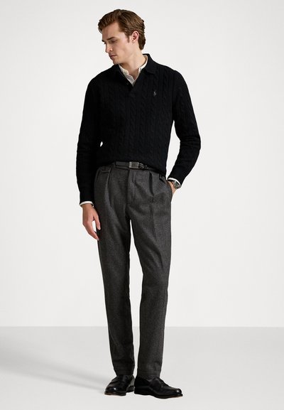 Pull en maille noir avec un col, associé à un pantalon plissé gris. Le modèle se tient sur un fond neutre, portant des chaussures noires.