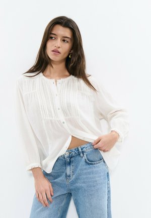 Young woman with long hair wearing white buttoned blouse and blue jeans, posing with one hand in pocket against white background.