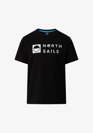 Garçon portant un t-shirt noir "North Sails", un short noir, des chaussettes blanches et des baskets blanches, debout les mains dans les poches contre un fond clair.