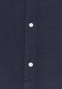 Marineblaues Hemd aus strukturiertem Stoff, mit einer Knopfleiste mit weißen Knöpfen und sichtbaren Nähten entlang der Kanten.