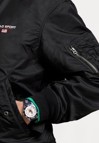 Svart nylon bombarjacka med ribbade muddar, utrustad med en dragkedjeficka och broderad logotyp. Armbandsur har ett blått och grönt silikonarmband.