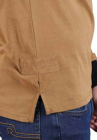 Chemise en coton marron avec un ourlet ventilé. Présente des coutures détaillées sur une pièce de tissu à l'arrière. Fini texturé.