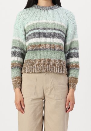 Femme portant un pull en tricot à rayures horizontales vertes, marron, blanches et grises, associé à un pantalon beige.