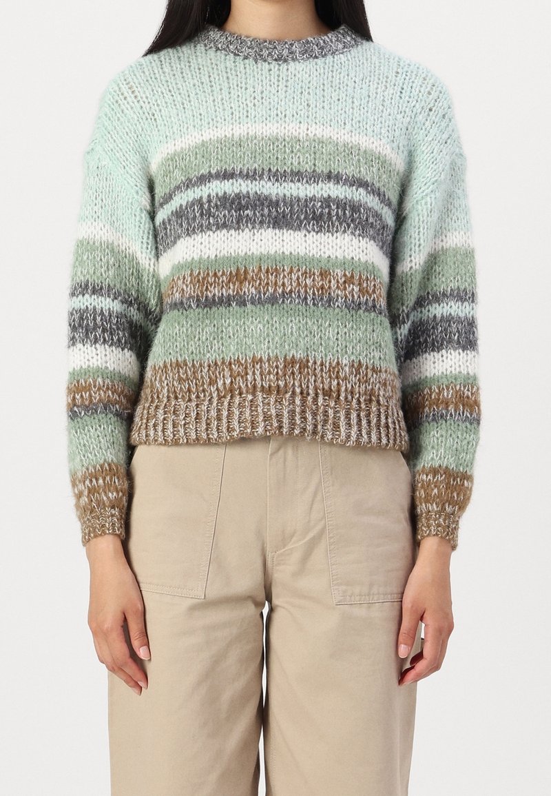 Femme portant un pull en tricot à rayures horizontales vertes, marron, blanches et grises, associé à un pantalon beige.