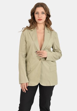 Donna con capelli castani mossi che indossa un blazer beige abbottonato in vita e pantaloni neri, in piedi davanti a uno sfondo grigio uniforme.