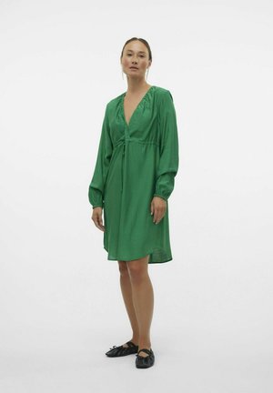 Abito verde a scollo a v con maniche lunghe e ampie e un nodo in vita. Tessuto morbido e lunghezza al ginocchio, abbinato a scarpe piatte nere.