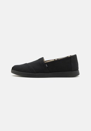 Melnas slip-on kurpes ar teksturētu kanvas augšējo daļu, ar zemu profilu un melnu gumijas zoli, kas izceļ minimālistisko dizainu un šuvju detaļas.