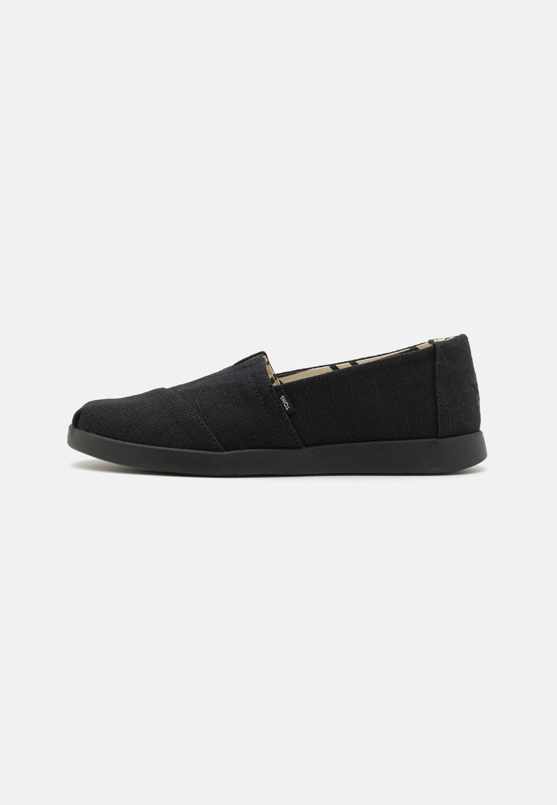 Black slip-on shoes with a textured canvas upper, featuring a low profile and a black rubber sole, showcasing minimalistic design and stitching details.