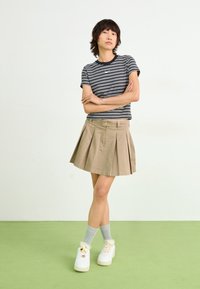 Persona de pie con los brazos cruzados, vestida con una camiseta a rayas blancas y negras, una falda plisada beige, calcetas grises, y zapatillas blancas sobre un suelo verde claro.