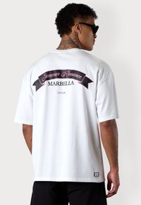 T-shirt blanc surdimensionné avec un graphisme noir et rose au dos portant l'inscription "Summer Romance MARBELLA", à manches courtes et coupe décontractée.
