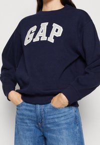 Sudadera azul marino con "GAP" bordado en color crema. Presenta un escote redondo y un corte holgado. Combinada con jeans de color azul claro.