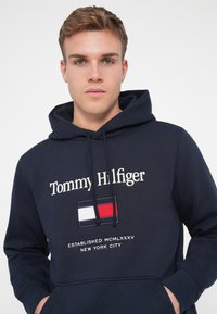 Felpa con cappuccio blu navy realizzata in tessuto morbido, con logo "Tommy Hilfiger", un emblematico striscione e testo ricamato al centro.