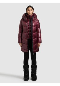 Sieviete, kas valkā spīdīgu tumši sarkanu ziemas mēteļa ar kapuci, melnas zeķubikses un melnus ziemas zābakus, stāvot pret platu fonu.