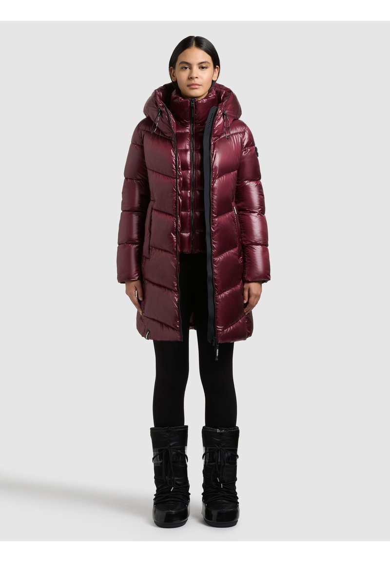 Sieviete, kas valkā spīdīgu tumši sarkanu ziemas mēteļa ar kapuci, melnas zeķubikses un melnus ziemas zābakus, stāvot pret platu fonu.