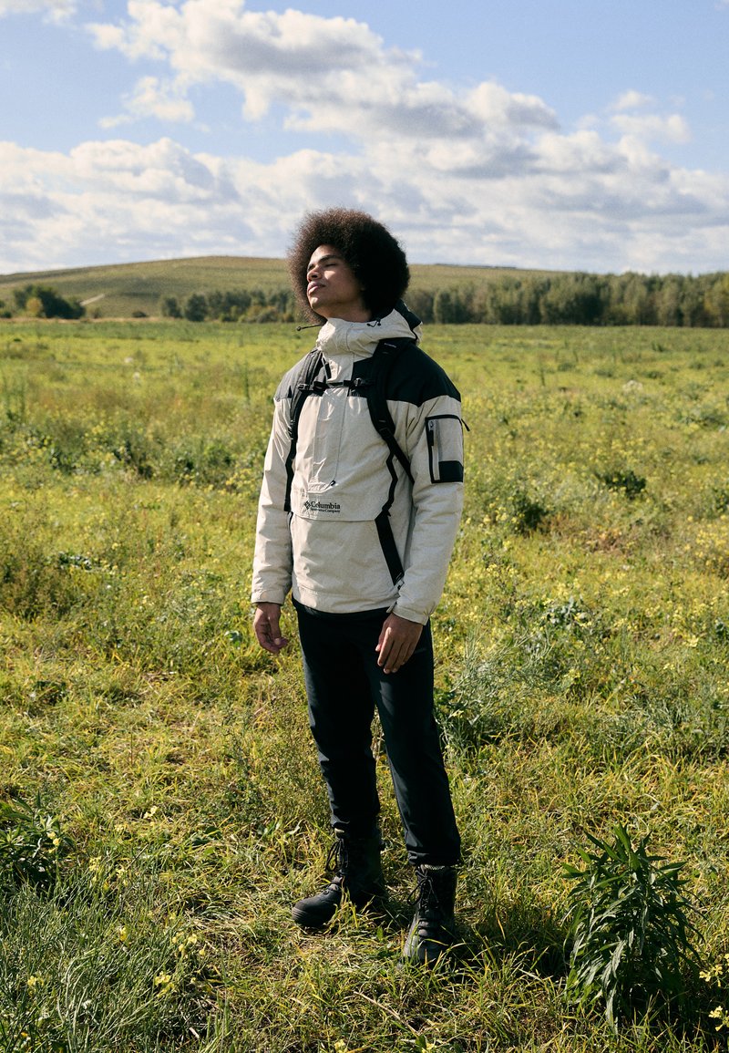 Beżowa kurtka z kapturem z czarnymi akcentami, szelkami i kieszenią na zamek, w połączeniu z dopasowanymi czarnymi spodniami i czarnymi butami z sznurowadłami, w trawiastym polu.