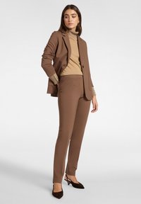 Bruin op maat gemaakt pak met een getailleerd blazer en slanke broek, gecombineerd met een beige coltrui. Gelaagde stof en knoopmanchetten voegen detail toe.