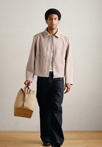 Jasnoróżowa wełniana kurtka zapinana na zamek, w połączeniu z ciemnymi dżinsami. Modelka niesie brązową torbę o usztywnionym kształcie i pojedynczym uchwycie.