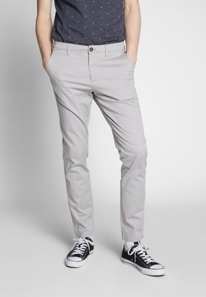 Mand iført lysegrå slimfit bukser, marineblå mønstret skjorte stukket ind, sort-hvide sneakers, stående med hænderne i lommerne.