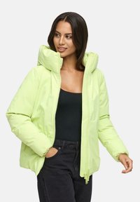 Vrouw met donker haar draagt een lichtgroene gewatteerde jas over een zwarte top en donkere spijkerbroek, met één hand in haar zak, kijkend zijwaarts.
