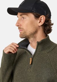 Dark green ribbed knit sweater with zippered collar, featuring a brown leather accent and a gray inner lining, paired with a black cap.