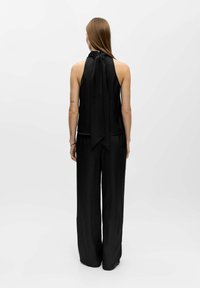 Schwarzer ärmelloser Jumpsuit mit hohem Hals und Schleifendetail am Rücken, aus glattem Stoff, mit weiten, fließenden Hosenbenen.
